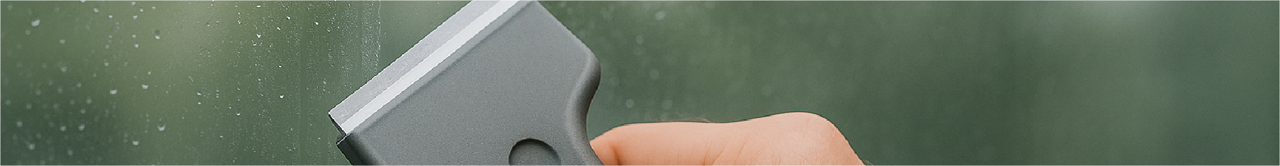 Hand holding a grey window scraper blade against a green background